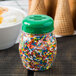 A Tablecraft green plastic slotted shaker top on a jar of sprinkles on a table with a bowl of ice cream.