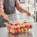 A person holding a Cambro clear plastic food storage container with a clear lid filled with red apples.