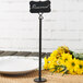 A black metal Tablecraft menu/card holder on a table with a sign next to a plate and flowers.