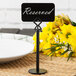A Tablecraft black metal menu holder on a table with a white sign.