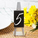 A Tablecraft stainless steel vertical card holder displaying a black table number sign on a table with yellow flowers.