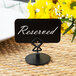 A black Tablecraft metal menu / card holder with a white sign reading "Reserved" on a table next to yellow flowers.
