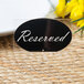 A black Tablecraft oval chalkboard label with white text on it next to yellow flowers on a table.