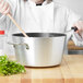 A woman in a white chef's coat uses a Vollrath Wear-Ever sauce pan with a black handle to stir food.