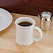 An Acopa ivory stoneware coffee mug filled with brown liquid on a wooden table.