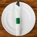 A white plate with a green customizable paper napkin band wrapped around a fork and knife.