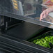 A hand opening a black Cambro Versa Well cover over a tray of greens on a salad bar.