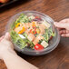 A hand holding a clear plastic container of salad with sauce in it.
