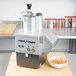 A Robot Coupe food processor on a table with a bowl of shredded carrots.