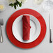 A 10 Strawberry Street red glass charger plate on a table with a white plate on top, a red napkin, and a fork and knife.