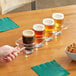 A hand holding an Acopa Acrylic Flight Paddle with Barbary Tasting Glasses filled with beer over a table with a bowl of pretzels.