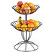 An American Metalcraft two-tiered wrought-iron basket filled with fruit on a table.