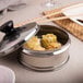 A stainless steel container of food with a white Choice 4" Perforated Steamer Paper lid on a table.