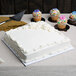 A white square cake on a white cake drum with cupcakes on a table.