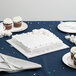 A white square cake on a table with cupcakes and a fork on a table with a white Enjay cake drum.