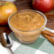 A glass bowl of Kime's sweetened cinnamon applesauce with a spoon and apples on a table.