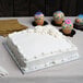 A white frosted cake on a silver square cake board with cupcakes on the table.