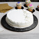 A white frosted cake on a black Enjay round cake drum on a table with cupcakes.
