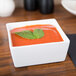 A white CAC china square bowl filled with tomato soup and a basil leaf on top.