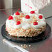 A cake with whipped cream and cherries on a black round cake board sitting on a table.