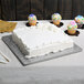 A white cake on a silver square cake drum on a table with cupcakes on top.