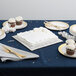 A table in a bakery display with a white cake on a silver square cake drum, surrounded by cupcakes and plates.