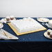 A white table with a white cake on a gold square cake drum.