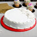 A white frosted cake on a red Enjay round cake drum on a table with cupcakes.