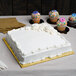 A white cake with frosting on a gold square cake drum with cupcakes on a table.