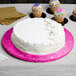 A white cake on a pink Enjay round cake drum on a table with cupcakes.