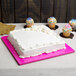 A white cake on a pink Enjay square cake drum on a table with cupcakes on a plate.