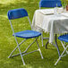 A table with a white tablecloth and blue Lancaster Table & Seating folding chairs on grass.