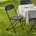 A Lancaster Table & Seating black folding chair on grass with a table set with chairs and a tablecloth.