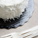 A white cake with white frosting on a black Enjay cake circle on a black plate with white plastic forks.