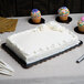 A white cake with frosting on a black Enjay laminated corrugated cake board on a table with cupcakes.