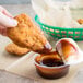 A hand holding a piece of fried chicken over a small container of Sweet Baby Ray's BBQ sauce.