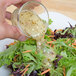 A hand pouring Ken's Foods Greek Vinaigrette with Feta Cheese and Black Olives dressing onto a salad.