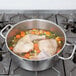 A Vollrath Optio brazier pot of chicken soup with vegetables on a stove.