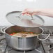 A hand with a metal handle holding a lid over a Vollrath Optio brazier pan filled with chicken soup.