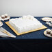 A white cake with gold and silver decorations on an Enjay gold square cake drum on a table.