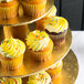 A tray of yellow frosted cupcakes with sprinkles displayed on a gold tiered stand.