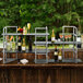 A 4 tier stainless steel riser with glass shelves holding wine glasses and bottles.