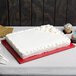A white cake with frosting on a red Enjay 1/2 sheet cake board on a table.