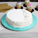 A white cake on a blue Enjay round cake drum with cupcakes on a table.