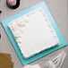 A blue Enjay square cake drum under a cake with white frosting on a white plate.
