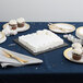 A table with a white cake on a black square cake drum, cupcakes, and plates with utensils.