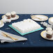 A blue square cake drum on a table with a white cake and utensils.
