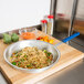 A Vollrath Wear-Ever aluminum fry pan filled with noodles and vegetables on a cutting board.