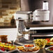 A KitchenAid food processor attachment on a table with a bowl of pineapple salad, a fruit tart, a bowl of salsa, and a plate of vegetables.