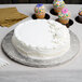 A white cake on a silver round cake drum on a table with cupcakes.